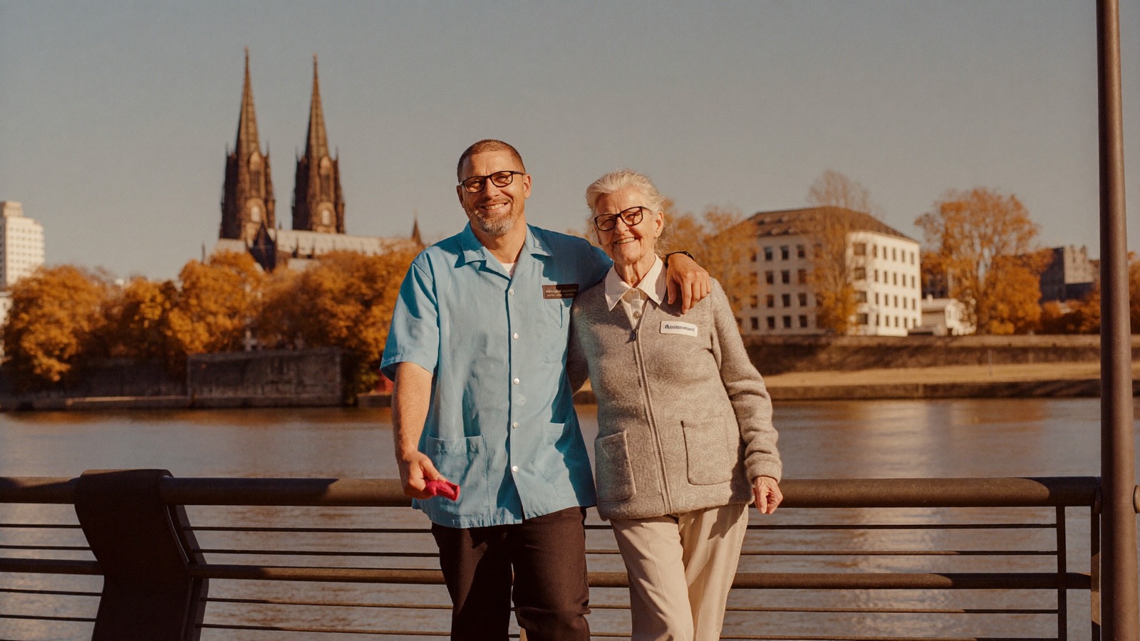 Betreuer spaziert Arm in Arm mit einer Seniorin an der Rheinpromenade in Köln – im Hintergrund der Kölner Dom