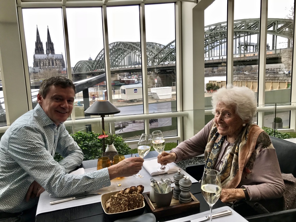 Inhaber Dieter Balling stößt mit einer Kundin an – im Hintergrund Kölner Dom und Hohenzollernbrücke